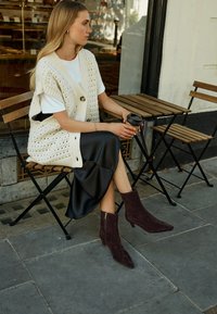 Oversized cream knit vest with large buttons layered over a white t-shirt; black satin skirt; burgundy suede ankle boots with zippers.