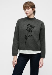 Jovem com cabelo curto, vestindo um suéter cinza escuro com um desenho de rosa contornado a preto, camisa branca por baixo e jeans escuros, pousando contra um fundo branco.