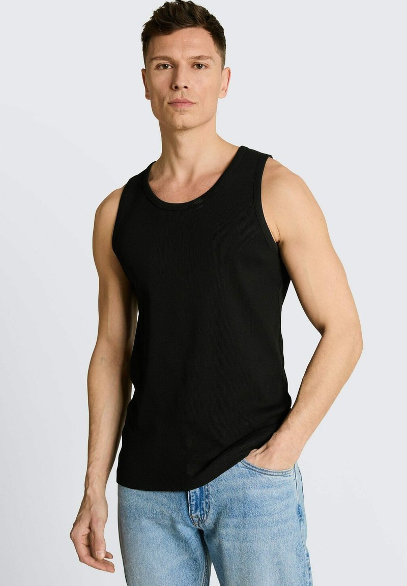 Jeune homme debout avec une main dans la poche, portant un débardeur noir sans manches et un jean bleu clair, fond neutre.