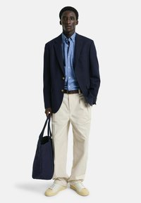 Mørkeblå blazer, lyseblå skjorte, beige bukser og hvite sneakers. Holder en mørk blå totebag. Enkelt design og avslappet passform.