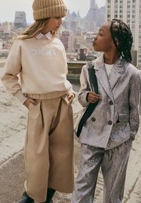 Deux enfants se tiennent sur un toit. L'un porte un sweat beige avec "DIONY" en strass et un pantalon large beige. L'autre porte un costume en sequins argentés.