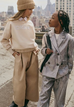Deux enfants se tiennent sur un toit. L'un porte un sweat beige avec "DIONY" en strass et un pantalon large beige. L'autre porte un costume en sequins argentés.