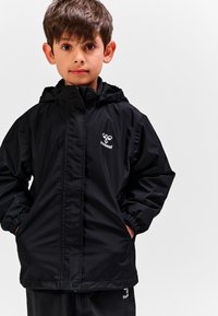 Czarna wodoodporna kurtka z kapturem, elastycznymi mankietami i dwoma przednimi kieszeniami. Na piersi znajduje się biały logo. Gładka, błyszcząca struktura.