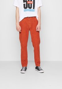 Pantalons en velours côtelé orange avec une coupe droite, dotés d'un bouton à l'avant et de poches avant, associés à des baskets noires et un t-shirt graphique.