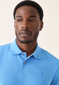 Világoskék póló, texturált anyagból, kétgombos patenttal és hímzett logóval a bal mellrészen.