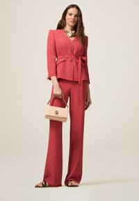 Różowy dopasowany garnitur z fasonem żakietu, szerokimi spodniami i paskiem w talii. W połączeniu z kremową torebką ozdobioną emblematem.