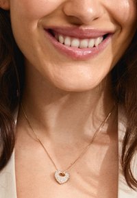 Collier en chaîne dorée avec un pendentif en forme de cœur, orné de cristaux transparents et d'un petit accent doré au centre.