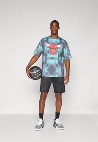 Basketball shorts i sort netstof, parret med en kortærmet, turkise tie-dye t-shirt med et rødt tyre-design. Holder en sort basketball.