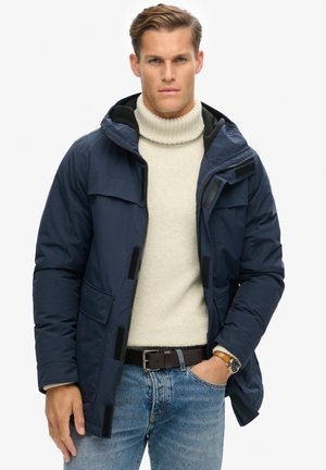 Marineblå pufferjakke med hætte, frontlynlås og Velcro-detaljer. Båret over en cremefarvet turtleneck, parret med lyseblå jeans.