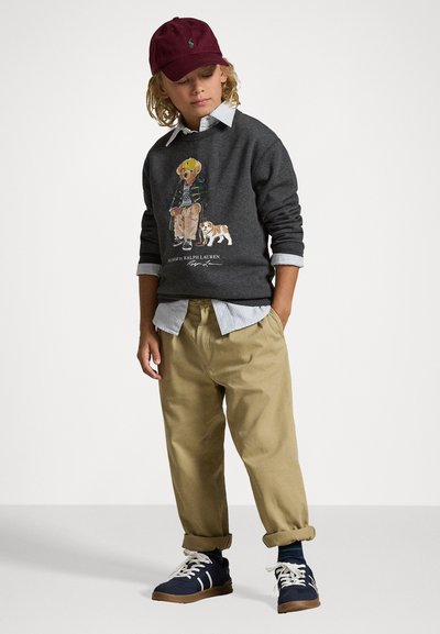 Siv sweatshirt iz bombažem z grafiko medveda, v kombinaciji z bež hlačami in mornarskimi supergami z belimi detajli. Burgundska kapa in svetlo modra srajca v plasteh.
