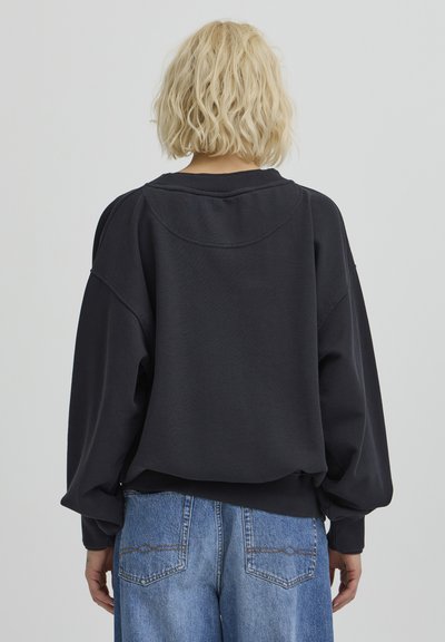 Sweatshirt noir oversized avec col et poignets côtelés. Le matériau semble doux avec une texture lisse. Porté avec un jean large bleu clair.
