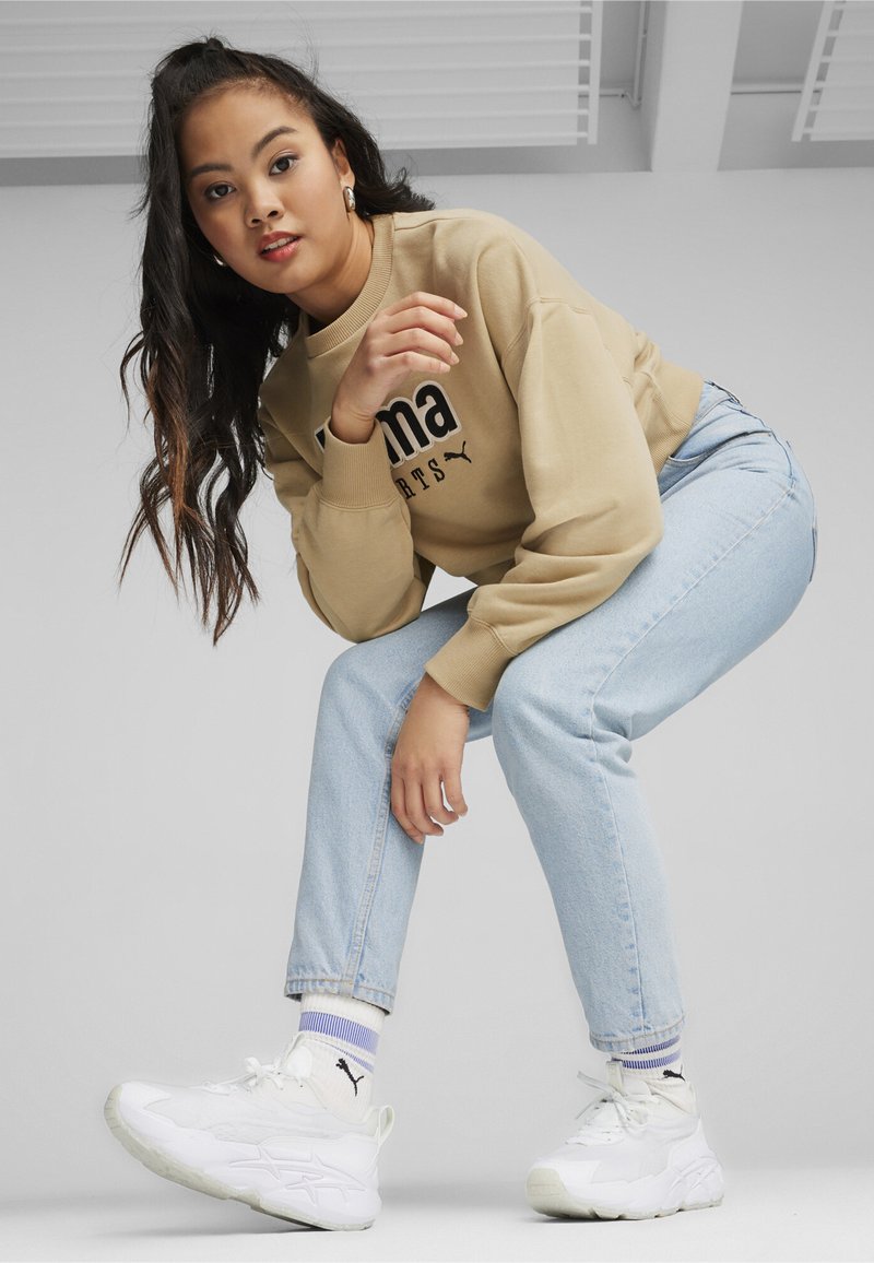Sweatshirt beige avec logo noir, jeans bleu clair et baskets blanches ; le mannequin est accroupi, montrant ses longs cheveux et des chaussettes avec accent violet.