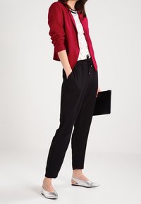 Blazer bordeaux sopra una maglietta di pizzo bianco, abbinato a pantaloni neri e ballerine argentate. L'outfit presenta un design elegante e contemporaneo, con diverse texture.