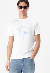 White cotton T-shirt featuring a light blue printed logo of a flag with "HACKETT" and "LONDON" beneath. The model wears denim jeans and sunglasses.
