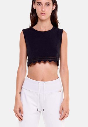 Donna che indossa un crop top nero senza maniche con orlo smerlato e pantaloni bianchi con coulisse, in piedi davanti a uno sfondo bianco semplice.
