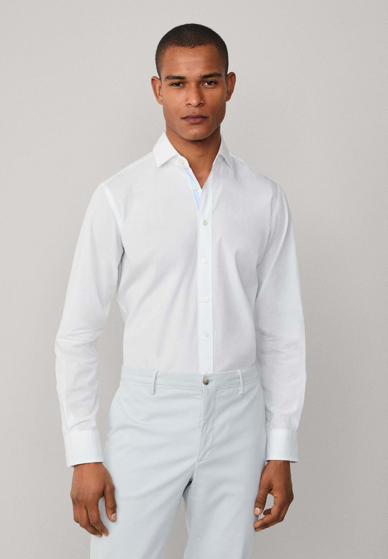 Camisa blanca de manga larga con cierre de botones, que presenta un acento en azul claro en el interior del cuello, combinada con pantalones azul claro.