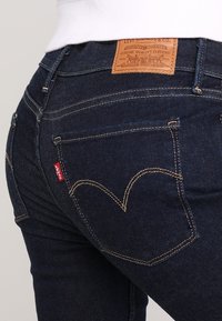 Jeans de mezclilla azul oscuro con un parche de cuero marrón con marca, costuras amarillas y un diseño de bolsillo trasero en forma de corazón.