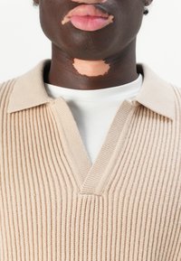 Maglione a coste beige con scollatura a V e colletto, indossato sopra una maglietta bianca a girocollo. Il tessuto appare morbido con una finitura testurizzata.