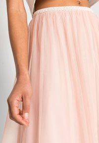 Jupon en tulle rose clair avec une taille ajustée, ornée d'une bordure en dentelle décorative. Le tissu a une texture douce et transparente.