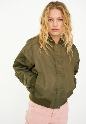 Chaqueta bomber verde oliva con puños y dobladillo acanalados, con cierre de cremallera y bolsillos laterales, combinada con pantalones rosas.