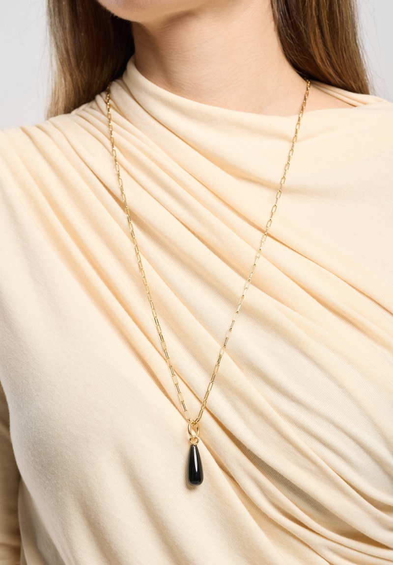 Goldkette mit einem länglichen schwarzen Anhänger. Die Kette hat ein Gliederdesign, und der Stoff fällt weich und in neutralen Tönen.