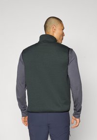 Gilet in pile verde scuro con colletto alto, design senza maniche e orlo elastico, indossato sopra una maglietta grigia a maniche lunghe. Stile semplice e funzionale.