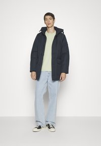 Navy puffer jacket with a hood, button closure, and front pocket, worn over a light green shirt and paired with light blue jeans and black sneakers.