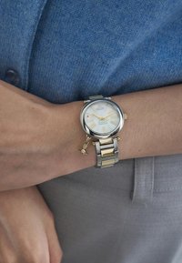 Ασημένιο και χρυσό μεταλλικό ρολόι με λευκή πρόσοψη, χρυσούς ρομαντικούς αριθμούς και μία μικρή γοητεία προσαρτημένη στο βραχιόλι. Φορεμένο σε καρπό.