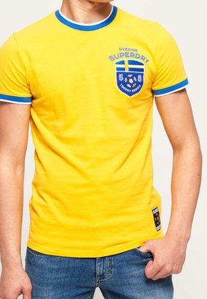 Mand iført en klar gul Superdry T-shirt med blå og hvide kanter og en blå logo med et svensk flag og tekst på brystet.