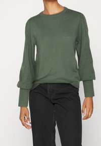 Pull en maille vert avec un col rond, des manches bouffantes et de subtils motifs en losange le long des bras. Porté avec un jean noir.