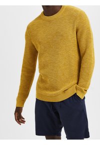 Gelber Strickpullover mit strukturiertem Muster, Rundhalsausschnitt und gerippten Bündchen, kombiniert mit dunkelblauen Shorts.