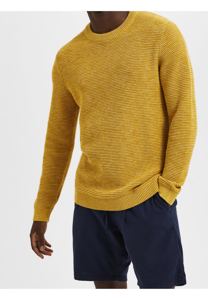 Gelber Strickpullover mit strukturiertem Muster, Rundhalsausschnitt und gerippten Bündchen, kombiniert mit dunkelblauen Shorts.