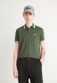 Grønn polo med to knapper, hvite detaljer på krage og ermer; brukt med svarte bukser og en grå caps med "BOSS"-logo.