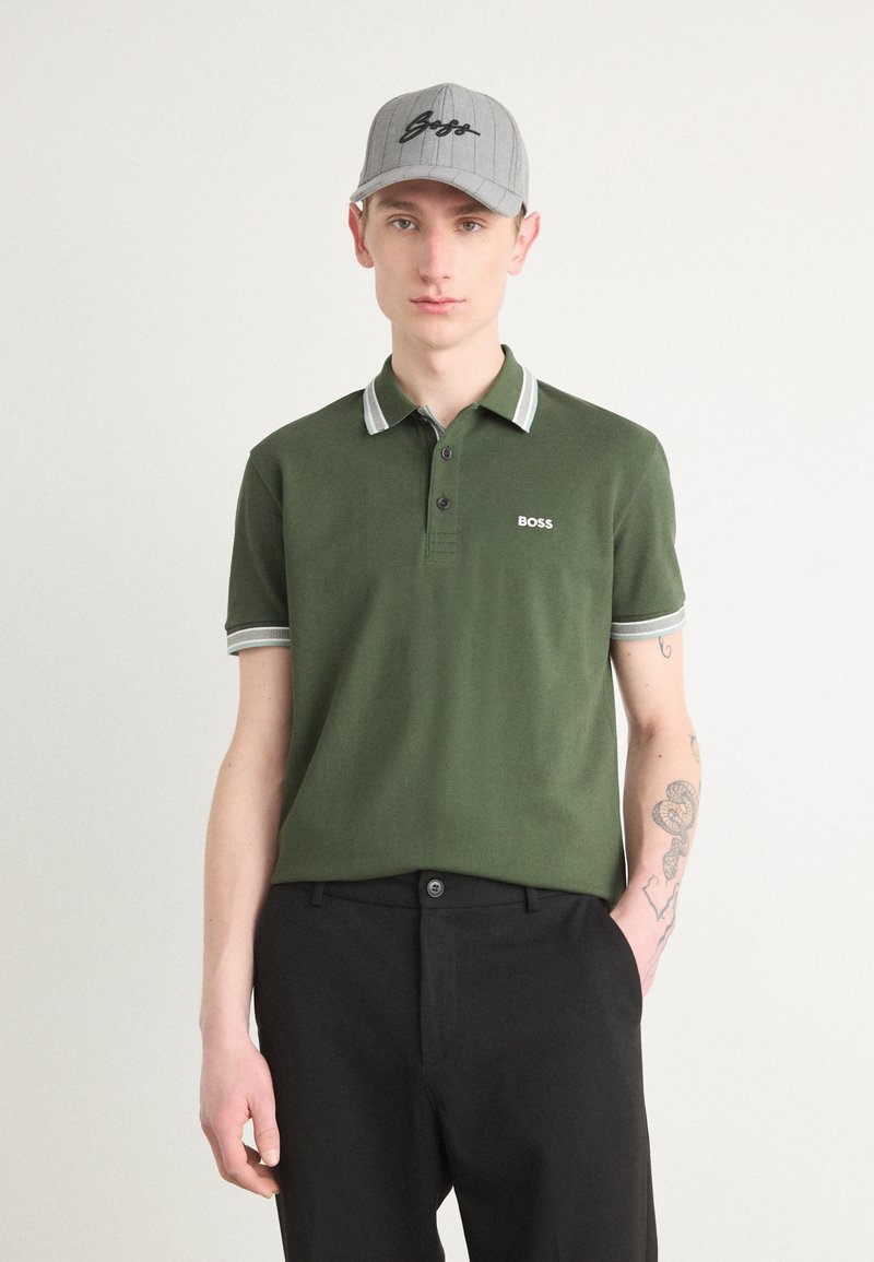 Grønn polo med to knapper, hvite detaljer på krage og ermer; brukt med svarte bukser og en grå caps med "BOSS"-logo.