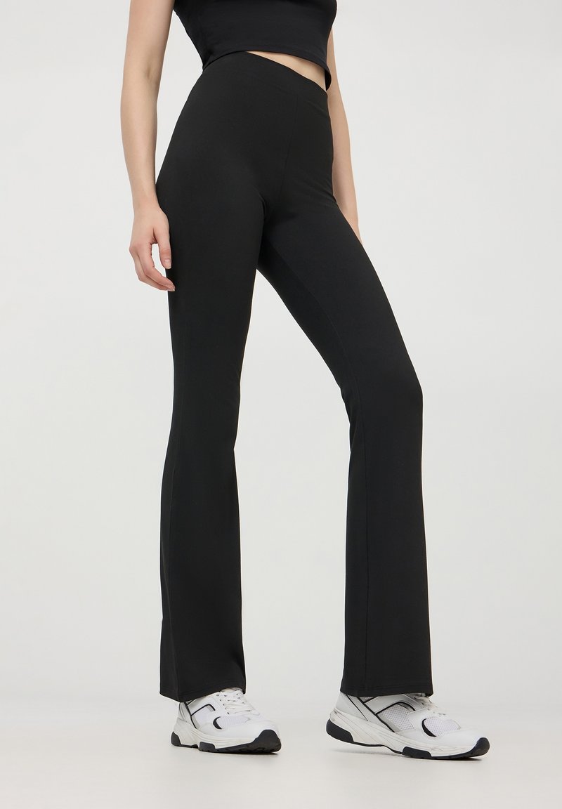 Personne portant un pantalon noir évasé taille haute et des baskets blanches, debout devant un fond clair uni.