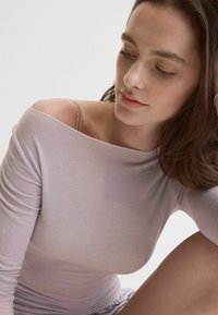 Mujer joven con cabello castaño largo que lleva una blusa lila clara con los hombros descubiertos, mirando hacia abajo con una expresión neutral sobre un fondo blanco.