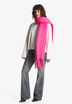 Fuksijasti volneni šal s frufrucami ovit okoli vratu modela. Uporabljen z belo jopo in razširjenimi denim hlačami, postavljen proti belemu ozadju.