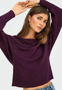 Haut long à manches ribblées violet avec un décolleté drapé, affichant une coupe décontractée et un tissu texturé. Porté avec un jean en denim bleu clair.