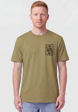 Mann mit roten Haaren, trägt olivgrünes T-Shirt mit kleinem schwarzem Joker-Kartengrafik auf der linken Brust, steht vor weißem Hintergrund.