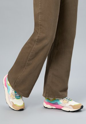 Personne portant un pantalon marron à jambes larges et des baskets colorées avec des touches de rose, bleu, vert et beige sur un fond gris.