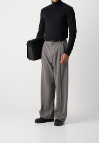 Schwarzer Rollkragenpullover, graue Plisseehose und schwarze Schuhe. Die Person hält eine schwarze Umhängetasche mit glatter Oberfläche.