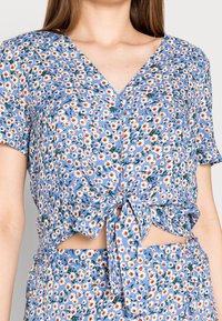 Crop top floral bleu clair avec un détail noué à l'avant. Comprend des manches courtes, un col en V, et un motif de marguerites et de feuilles vertes.