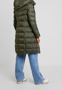 Mørkegrøn pufferjakke med hætte, der har et quiltet design og elastiske manchetter. Sammen med lyseblå vide jeans og beige sneakers.