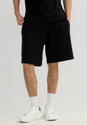 Hombre con pantalones cortos negros, calcetas altas blancas y zapatillas blancas de pie con las manos en los bolsillos.