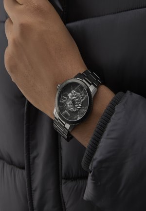 Černé metalické hodinky s texturovaným černým ciferníkem s designem růže, stříbrnými akcenty a náramkem s propojenými články.