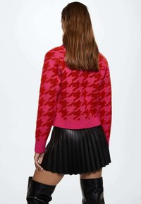 Femme aux longs cheveux bruns portant un pull en pied-de-poule rouge et rose, une jupe plissée noire et des bottes noires au-dessus du genou, vue de dos.