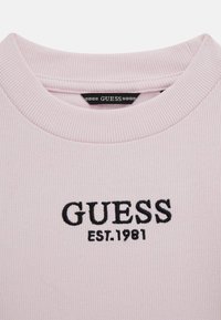 Világos rózsaszín bordázott pulóver kerek nyakkal. Elöl feltűnően fekete hímzett "GUESS EST.1981" felirattal rendelkezik.