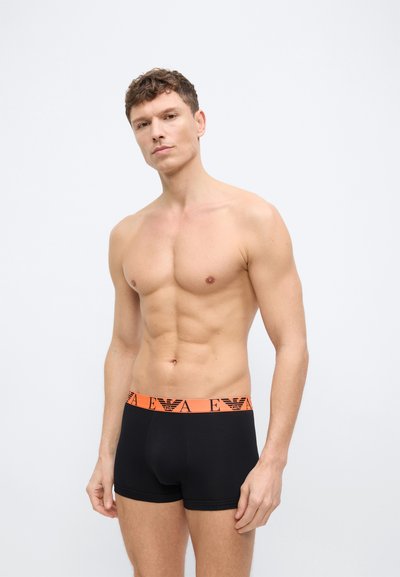 Ανδρας χωρίς μπλούζα με κοντά σγουρά μαλλιά, φορώντας μαύρα μποξεράκια με πορτοκαλί μέση που φέρει λογότυπο, στέκεται μπροστά σε απλό λευκό φόντο.