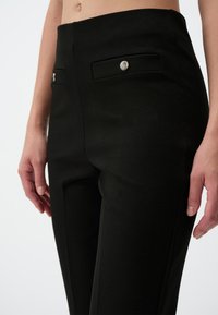 Zwarte op maat gemaakte broek heeft een flat front met twee kleine, decoratieve zakken en hardware in zilverkleur. De stof heeft een gladde, matte textuur.