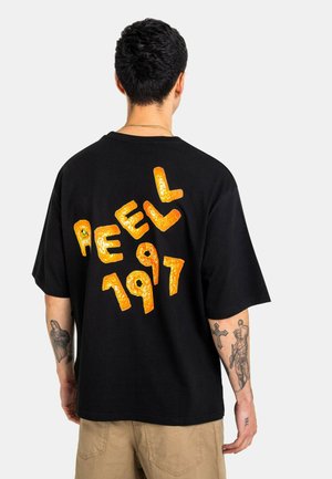 Man with short dark hair wears black oversized t-shirt with orange textured lettering "PEELL 1997" on back, tan pants, and arm tattoos.